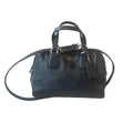 A black pebbled leather Coach handbag with gold-tone hardware, resting on a light wood floor. The bag features dual top handles, a detachable shoulder strap, a hanging leather tag, and a small gold Coach logo plaque on the front.