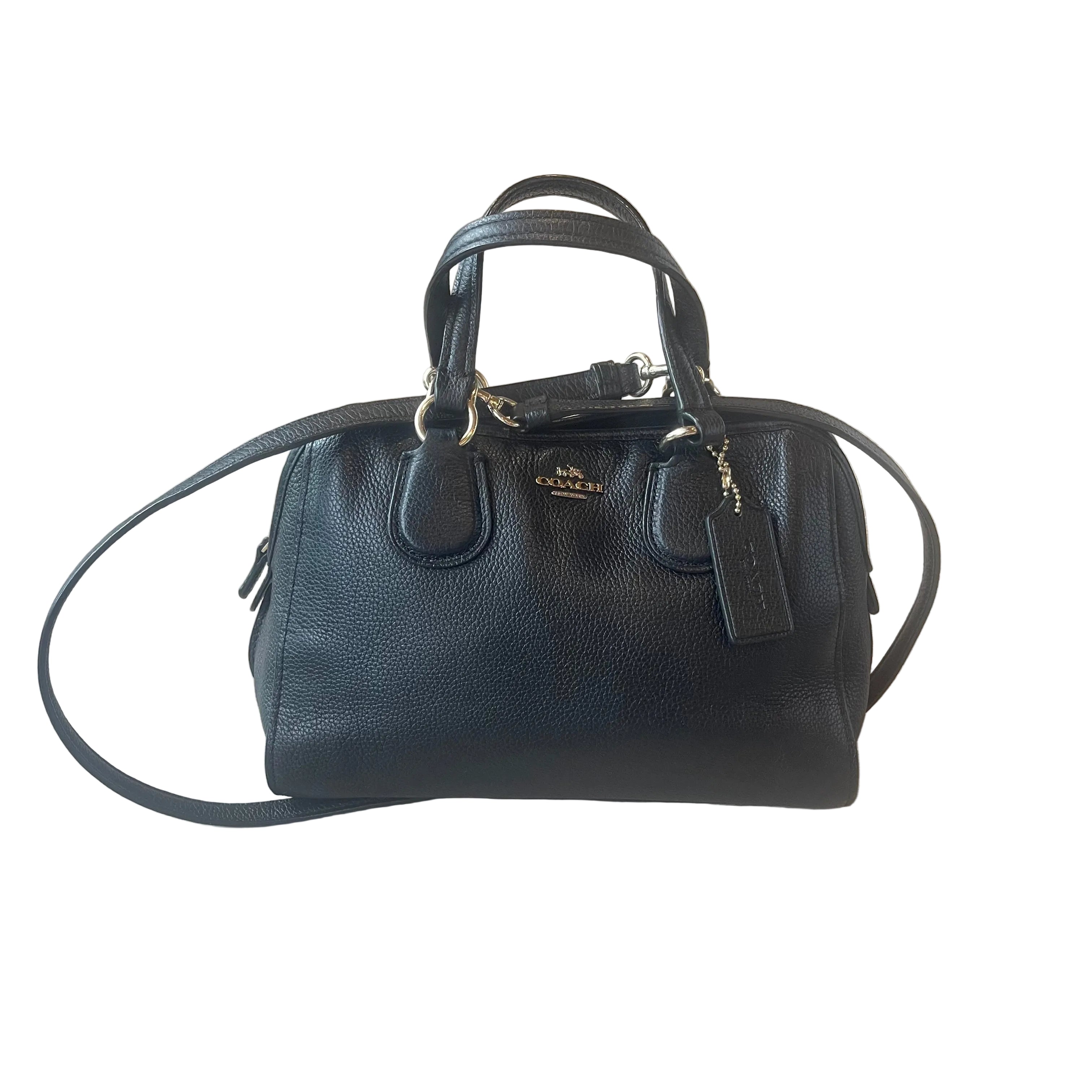A black pebbled leather Coach handbag with gold-tone hardware, resting on a light wood floor. The bag features dual top handles, a detachable shoulder strap, a hanging leather tag, and a small gold Coach logo plaque on the front.
