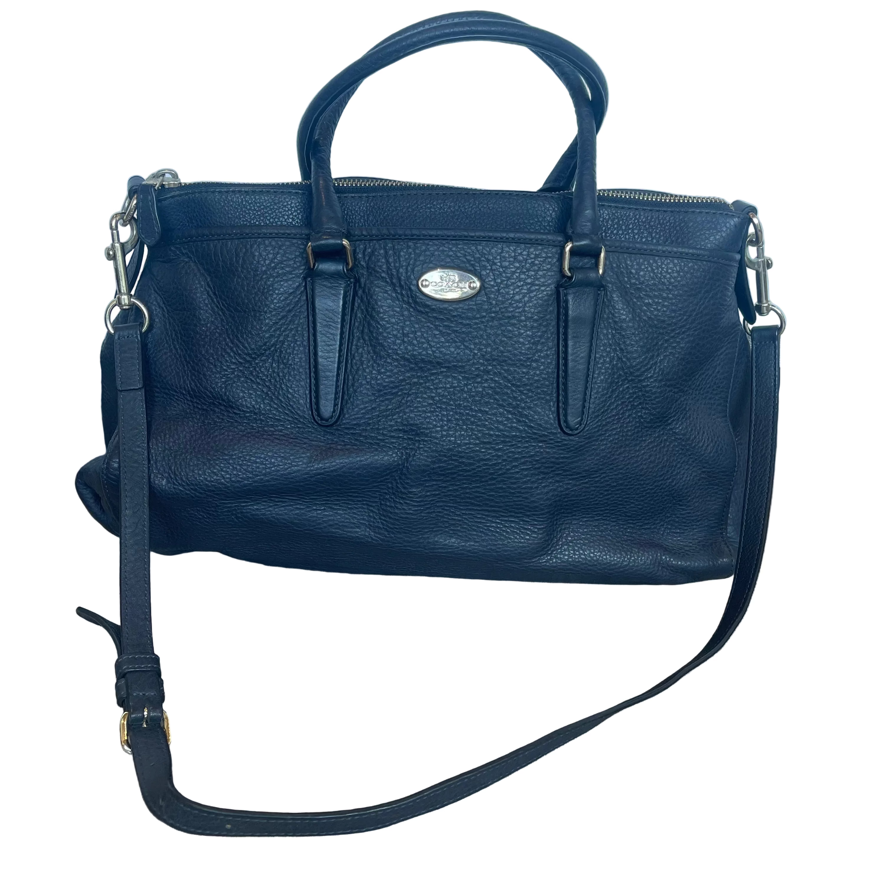 A black pebbled leather handbag with double top handles, a detachable shoulder strap, and a silver-tone oval logo plaque on the front, displayed on a wood-look floor against a light blue fabric backdrop.