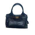 A black leather handbag with double top handles and a wide decorative belt featuring a gold-tone circular buckle, shown on a wood floor against a white fabric backdrop.