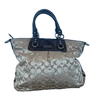 A Coach handbag in beige and brown signature monogram canvas with dark brown leather handles and trim, resting on a wooden floor against a light blue fabric backdrop.