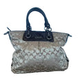 A Coach handbag in beige and brown signature monogram canvas with dark brown leather handles and trim, resting on a wooden floor against a light blue fabric backdrop.