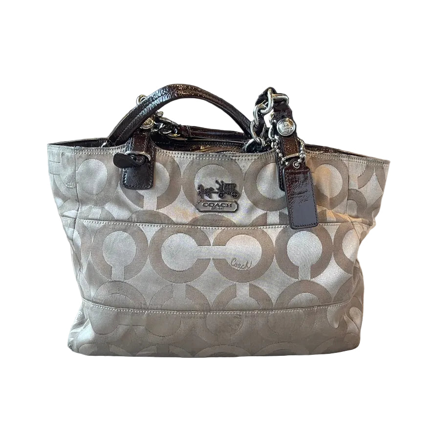 A Coach tote bag in signature beige and taupe monogram canvas with dark brown leather handles and a silver-tone chain detail, resting on a light wood floor.