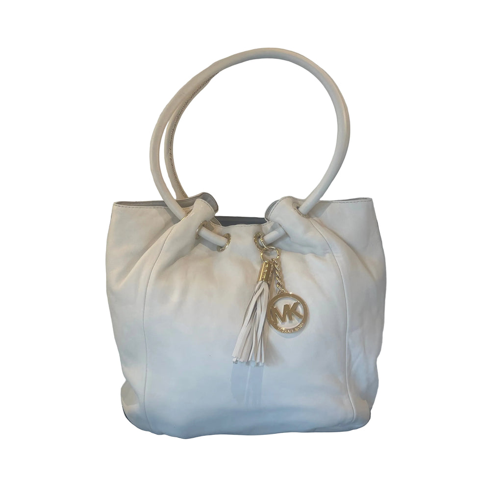 A cream-colored Michael Kors leather bucket bag with a drawstring top, dual top handles, and a gold-tone MK logo charm with a tassel, resting on a black chair against a white wall.