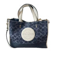 A Coach tote bag in navy and beige signature monogram canvas, featuring a large beige oval logo patch on the front, dual top handles, and a shoulder strap, resting on a light wood floor.