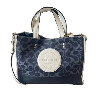 A Coach tote bag in navy and beige signature monogram canvas, featuring a large beige oval logo patch on the front, dual top handles, and a shoulder strap, resting on a light wood floor.